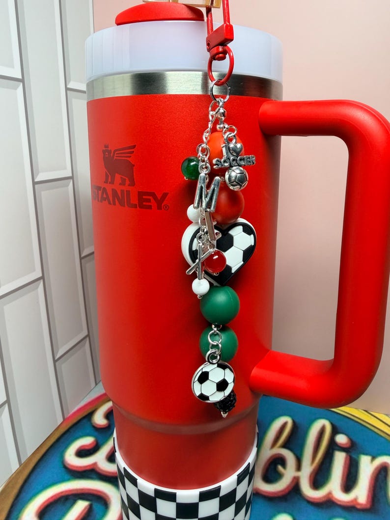 Puede incluir: Un vaso Stanley de color rojo brillante con asa y tapa blanca, que presenta un dije decorativo sujeto en la parte superior. El dije incluye una letra M plateada, un dije de bal&oacute;n de f&uacute;tbol, una cuenta en forma de coraz&oacute;n y cuentas verdes, rojas y blancas.