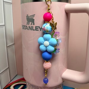 May include: A decorative beaded keychain featuring a gold-tone lobster clasp, a bright pink silicone bead, a blue floral patterned bead, a large light blue flower bead, a matte blue bead, a textured pink bead, and a small translucent purple bead.