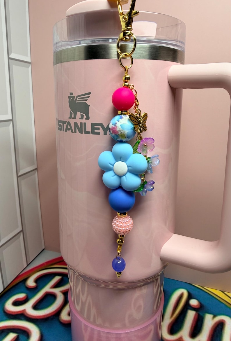 May include: A decorative beaded keychain featuring a gold-tone lobster clasp, a bright pink silicone bead, a blue floral patterned bead, a large light blue flower bead, a matte blue bead, a textured pink bead, and a small translucent purple bead.