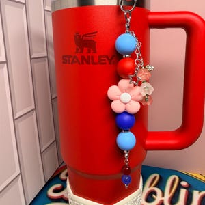 Op de afbeelding: Een rode Stanley-beker met een handvat en een decoratieve bedel. De bedel heeft blauwe, rode en roze kralen, samen met ster- en bloemaccenten. De beker heeft een witte, kanten basis.