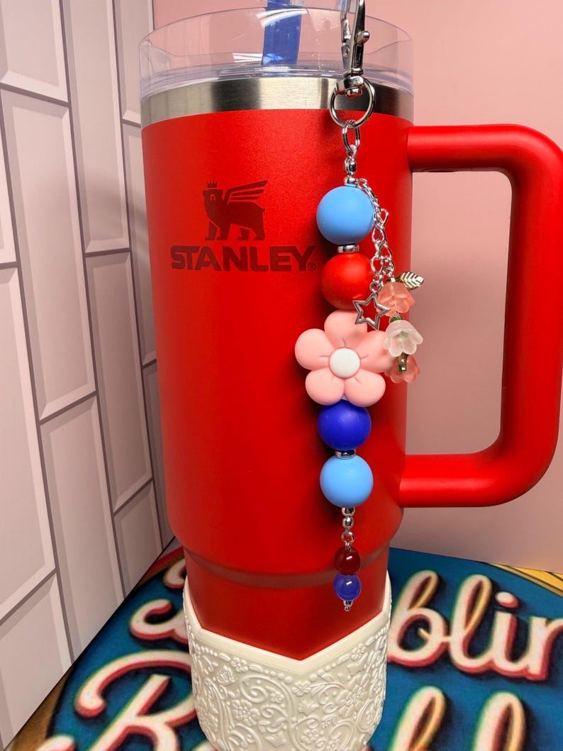 Op de afbeelding: Een rode Stanley-beker met een handvat en een decoratieve bedel. De bedel heeft blauwe, rode en roze kralen, samen met ster- en bloemaccenten. De beker heeft een witte, kanten basis.