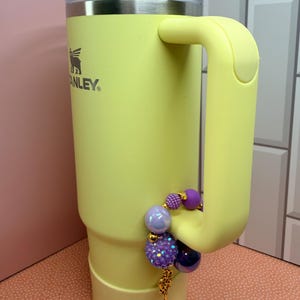 Op de afbeelding: Een lichtgele Stanley-beker met een handvat en een doorzichtig deksel. Het is versierd met een bedelarmband met paarse en iriserende kralen en een goudkleurige bedel. Het Stanley-logo is zichtbaar aan de zijkant.