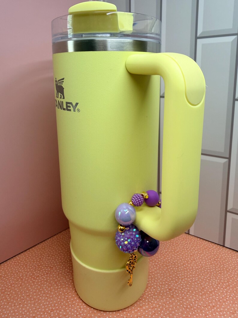 Op de afbeelding: Een lichtgele Stanley-beker met een handvat en een doorzichtig deksel. Het is versierd met een bedelarmband met paarse en iriserende kralen en een goudkleurige bedel. Het Stanley-logo is zichtbaar aan de zijkant.