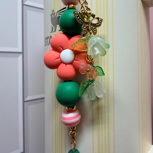 May include: A decorative charm featuring a central coral flower bead, flanked by green and pink-striped spherical beads. It includes a gold-toned butterfly charm and translucent green and pink bell-shaped flower beads on a gold metal chain.