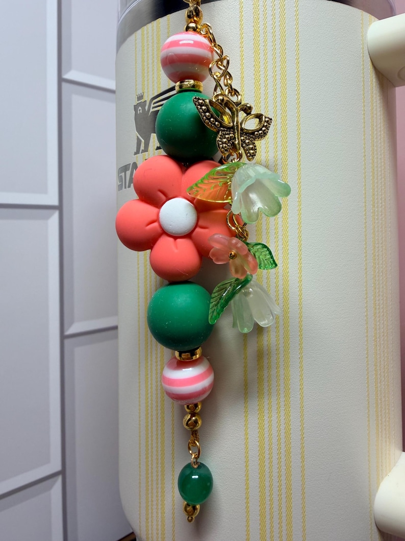 May include: A decorative charm featuring a central coral flower bead, flanked by green and pink-striped spherical beads. It includes a gold-toned butterfly charm and translucent green and pink bell-shaped flower beads on a gold metal chain.
