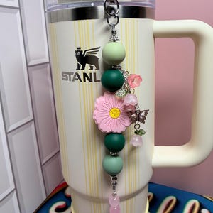 May include: A decorative tumbler charm featuring a silver lobster clasp, matte silicone beads in shades of sage and forest green, a large pink flower bead with a yellow center, and silver-toned charms including a butterfly and small pink flowers.