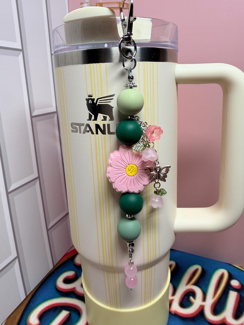 May include: A decorative tumbler charm featuring a silver lobster clasp, matte silicone beads in shades of sage and forest green, a large pink flower bead with a yellow center, and silver-toned charms including a butterfly and small pink flowers.