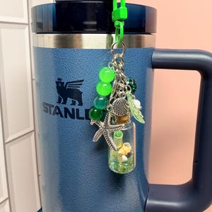 May include: A blue Stanley tumbler with a handle and a straw, featuring a decorative keychain attached to the lid. The keychain includes green beads, a silver starfish charm, a seashell charm, and a small glass bottle filled with tiny shells.