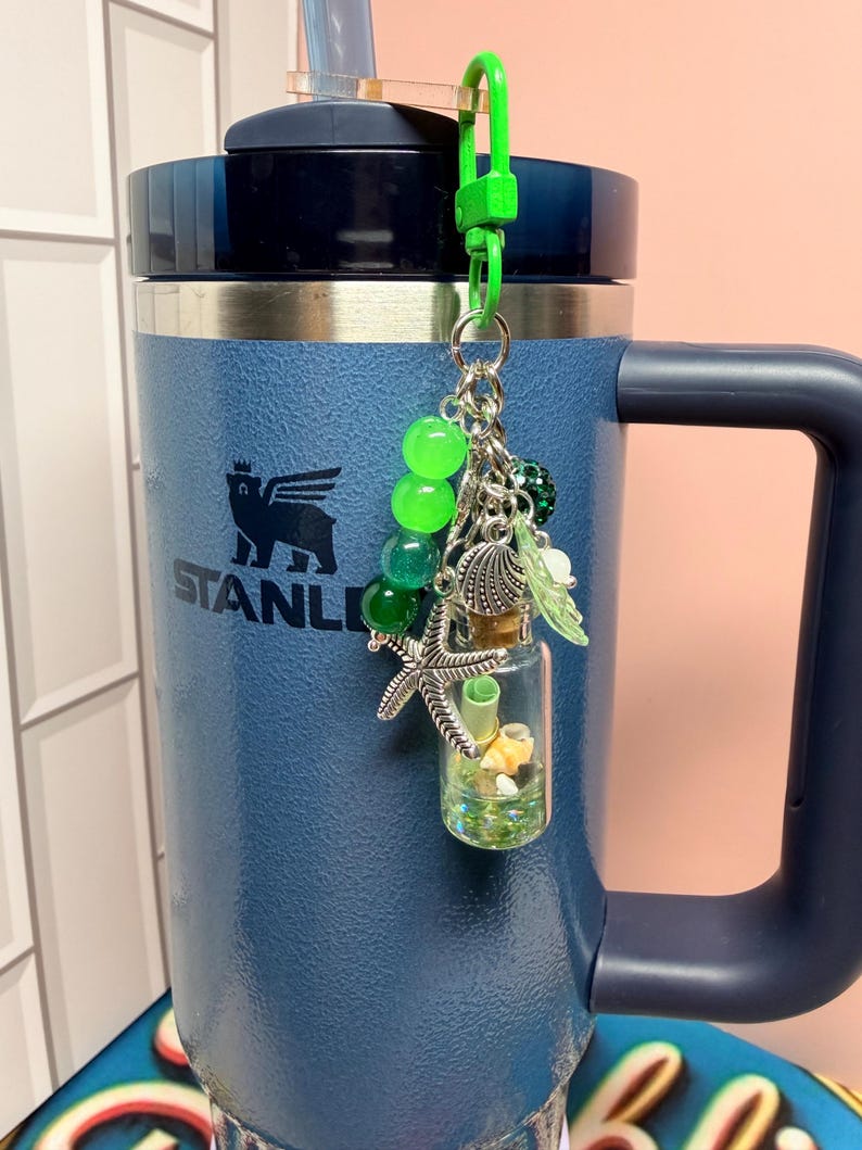 May include: A blue Stanley tumbler with a handle and a straw, featuring a decorative keychain attached to the lid. The keychain includes green beads, a silver starfish charm, a seashell charm, and a small glass bottle filled with tiny shells.