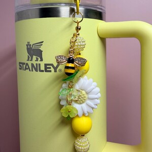 May include: A yellow Stanley tumbler with a decorative charm attached to the handle. The charm features a bee with rhinestone wings, a large white daisy, green leaf accents, and various yellow and white beads on a gold-toned metal chain.