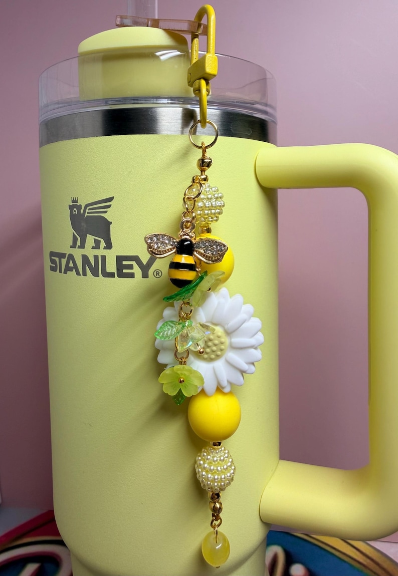 May include: A yellow Stanley tumbler with a decorative charm attached to the handle. The charm features a bee with rhinestone wings, a large white daisy, green leaf accents, and various yellow and white beads on a gold-toned metal chain.