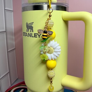 May include: A yellow Stanley tumbler with a decorative charm attached to the handle. The charm features a gold-toned bee with crystal wings, a white daisy flower, translucent green leaf beads, and several yellow and pearl-textured round beads.