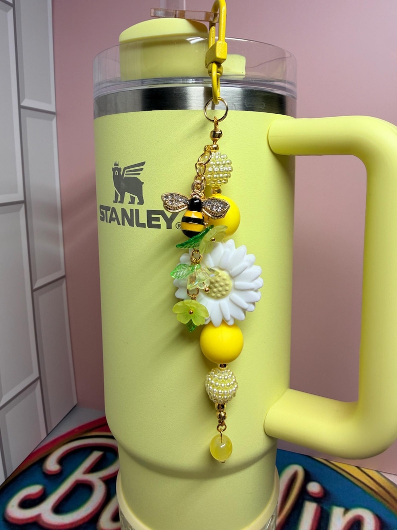 May include: A yellow Stanley tumbler with a decorative charm attached to the handle. The charm features a gold-toned bee with crystal wings, a white daisy flower, translucent green leaf beads, and several yellow and pearl-textured round beads.