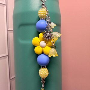 May include: A beaded charm hanging from a teal bottle, featuring a large yellow flower bead, smooth light blue spheres, textured yellow beads, a silver sun charm, and translucent yellow bell-shaped flower pendants on a silver metal chain.