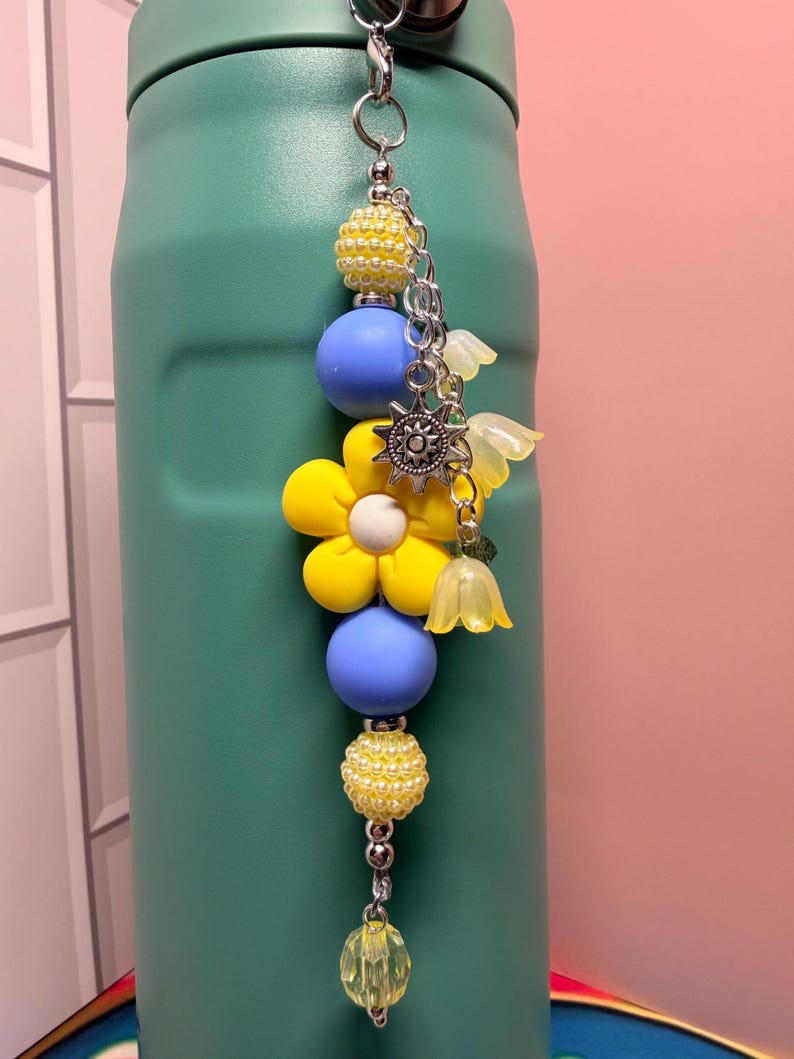May include: A beaded charm hanging from a teal bottle, featuring a large yellow flower bead, smooth light blue spheres, textured yellow beads, a silver sun charm, and translucent yellow bell-shaped flower pendants on a silver metal chain.