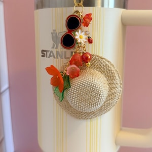 May include: A decorative charm with a gold clasp, featuring a miniature straw hat, red sunglasses, and floral accents. The charm is attached to a cream-coloured tumbler with the word "STANLEY" visible.
