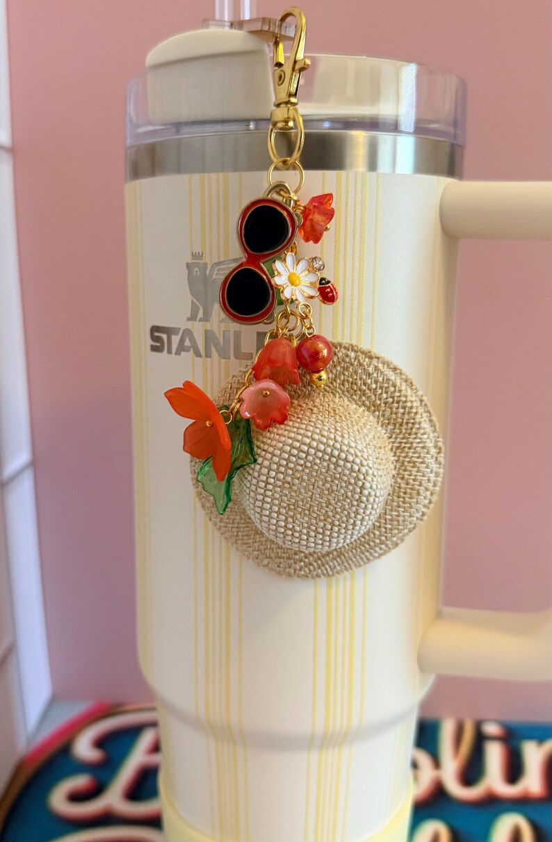 May include: A decorative charm with a gold clasp, featuring a miniature straw hat, red sunglasses, and floral accents. The charm is attached to a cream-coloured tumbler with the word "STANLEY" visible.