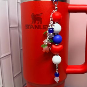 Op de afbeelding: Een rode Stanley-beker met een blauwe rietje en een decoratieve bedel. De bedel heeft rode, witte en blauwe kralen, samen met kleine bloemaccenten. De beker heeft een handvat en een witte decoratieve basis.