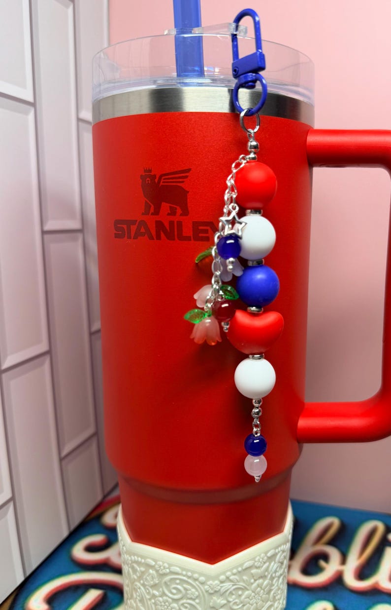 Op de afbeelding: Een rode Stanley-beker met een blauwe rietje en een decoratieve bedel. De bedel heeft rode, witte en blauwe kralen, samen met kleine bloemaccenten. De beker heeft een handvat en een witte decoratieve basis.