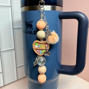 K&ouml;nnte beinhalten: Ein blauer isolierter Becher mit Griff und dekorativem Anh&auml;nger. Der Anh&auml;nger hat pfirsich- und cremefarbene Perlen, ein herzf&ouml;rmiges B&uuml;cherregal, einen kleinen Pompon und silberne Charms. Der Becher hat das "STANLEY"-Logo.