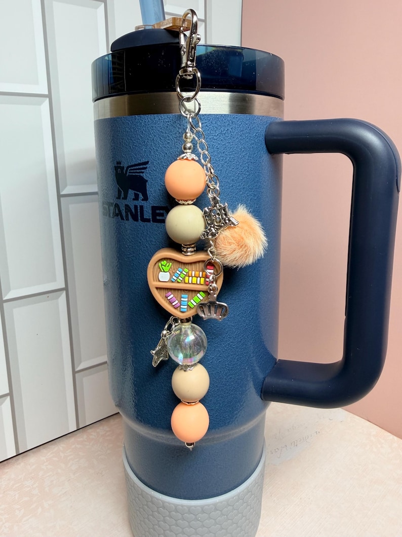 K&ouml;nnte beinhalten: Ein blauer isolierter Becher mit Griff und dekorativem Anh&auml;nger. Der Anh&auml;nger hat pfirsich- und cremefarbene Perlen, ein herzf&ouml;rmiges B&uuml;cherregal, einen kleinen Pompon und silberne Charms. Der Becher hat das "STANLEY"-Logo.