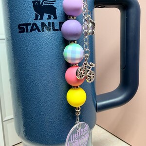 May include: A colourful beaded charm with a bicycle charm and a clear acrylic tag that says "Hello Spring". The charm features beads in purple, pink, yellow, and green. The charm is attached to a blue insulated tumbler.