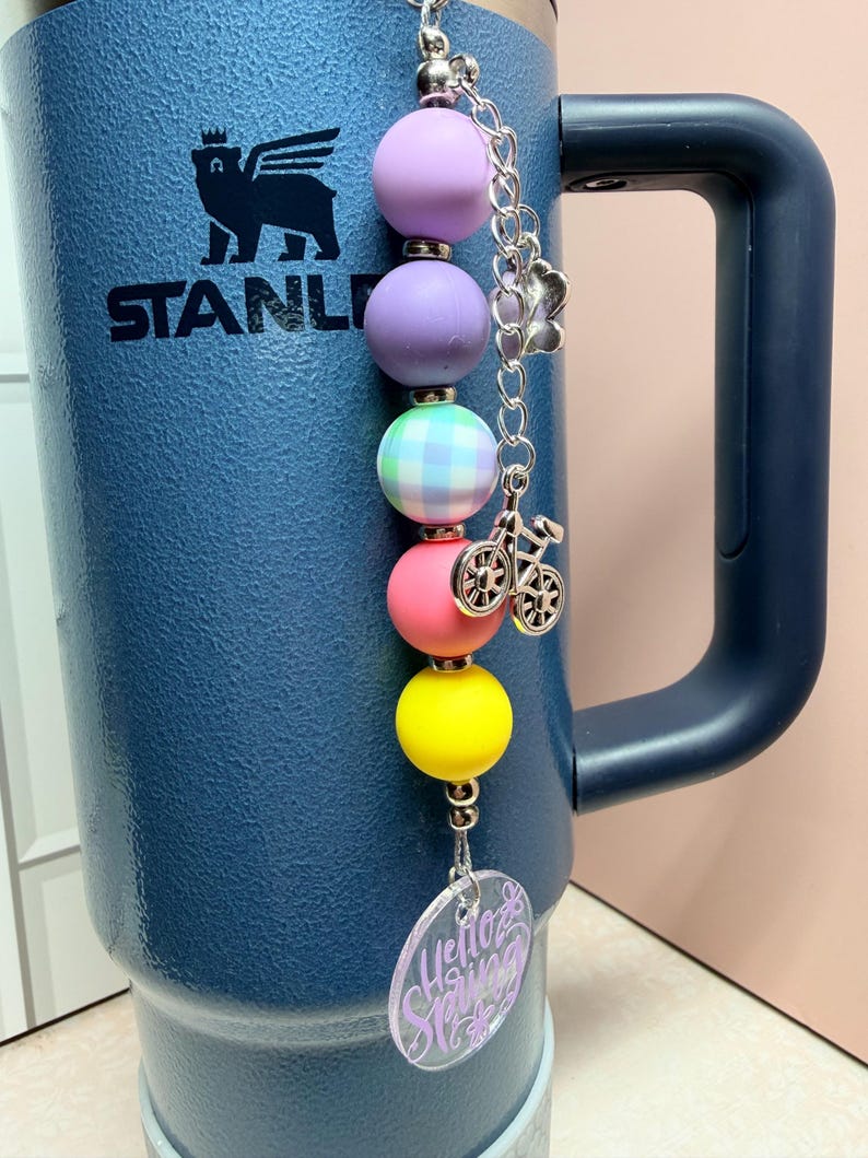 May include: A colourful beaded charm with a bicycle charm and a clear acrylic tag that says "Hello Spring". The charm features beads in purple, pink, yellow, and green. The charm is attached to a blue insulated tumbler.