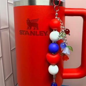 Op de afbeelding: Een rode Stanley-beker met bijpassende handgreep en een blauw rietje. Een decoratieve bedel met rode, witte en blauwe kralen en bloemaccenten hangt aan het deksel. Het Stanley-logo is zichtbaar.