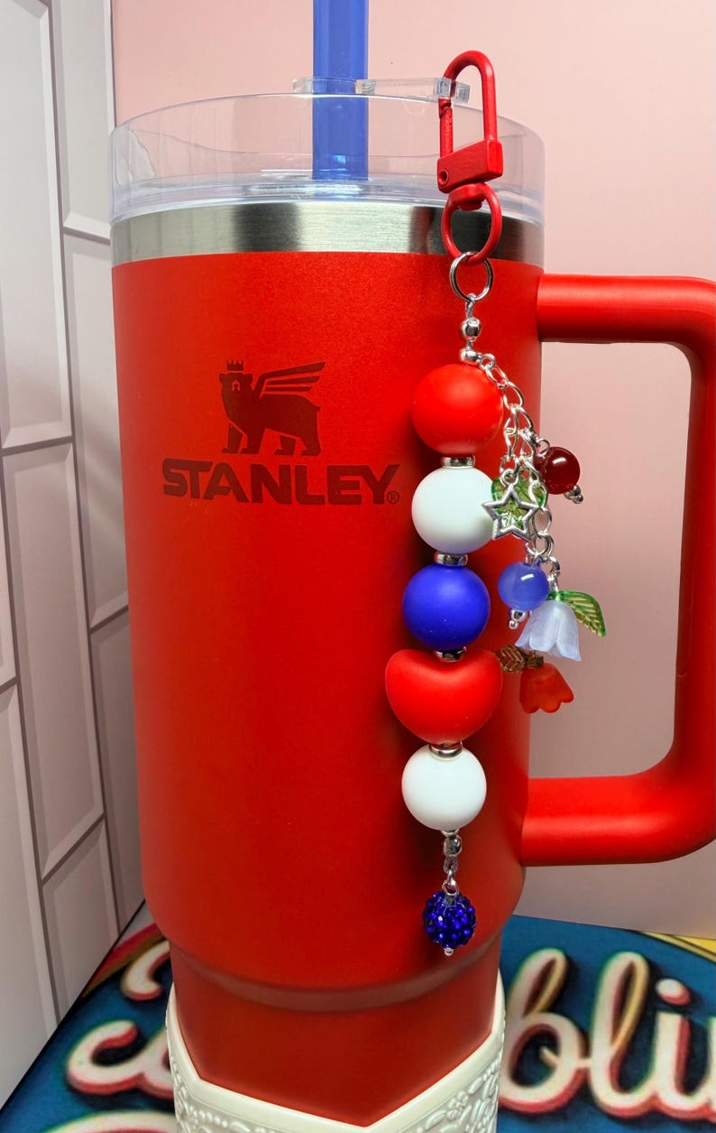 Op de afbeelding: Een rode Stanley-beker met bijpassende handgreep en een blauw rietje. Een decoratieve bedel met rode, witte en blauwe kralen en bloemaccenten hangt aan het deksel. Het Stanley-logo is zichtbaar.