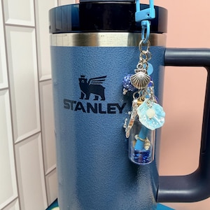 May include: A blue Stanley tumbler with a handle and a straw, featuring a decorative keychain attached to the lid. The keychain includes a small clear glass vial filled with blue glitter, a silver seashell charm, and a light blue flower-shaped bead.