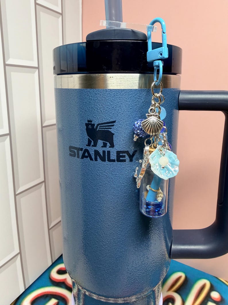 May include: A blue Stanley tumbler with a handle and a straw, featuring a decorative keychain attached to the lid. The keychain includes a small clear glass vial filled with blue glitter, a silver seashell charm, and a light blue flower-shaped bead.