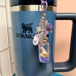 May include: A blue insulated tumbler with a handle and a black lid, featuring a hanging charm accessory. The charm includes a small glass bottle with a cork stopper containing a rolled paper scroll, a seahorse, a starfish, and purple beads.