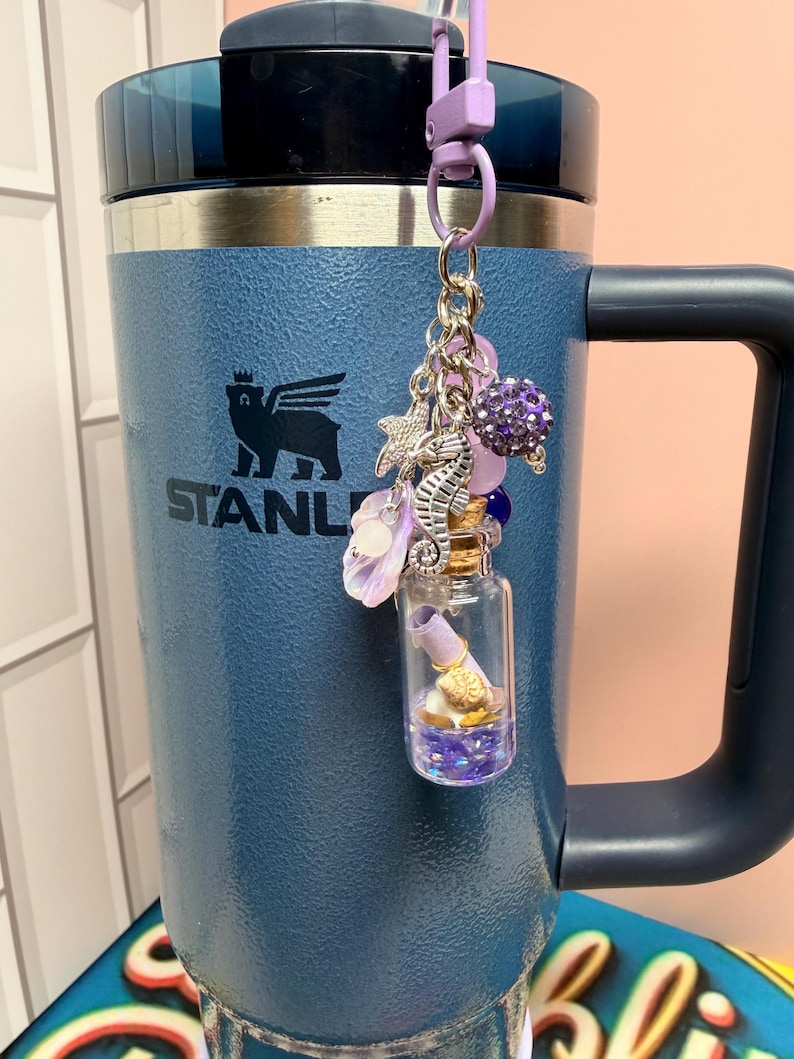 May include: A blue insulated tumbler with a handle and a black lid, featuring a hanging charm accessory. The charm includes a small glass bottle with a cork stopper containing a rolled paper scroll, a seahorse, a starfish, and purple beads.