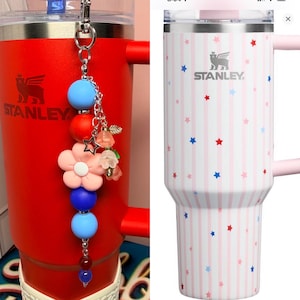 Op de afbeelding: Twee Stanley-bekers. E&eacute;n is rood met een decoratieve bedel. De bedel heeft blauwe, rode en roze kralen, een ster en bloemen. De andere is wit met roze strepen en blauwe en rode sterren. Beide hebben het Stanley-logo.