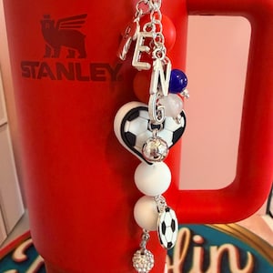 Puede incluir: Un vaso Stanley de color rojo brillante con asa tiene un llavero decorativo colgando de la parte superior. El llavero incluye dijes de letras plateadas que forman la palabra TENG, un dije de coraz&oacute;n con forma de bal&oacute;n de f&uacute;tbol, cuentas redondas blancas, naranjas y azules, y un peque&ntilde;o dije de bal&oacute;n de f&uacute;tbol.