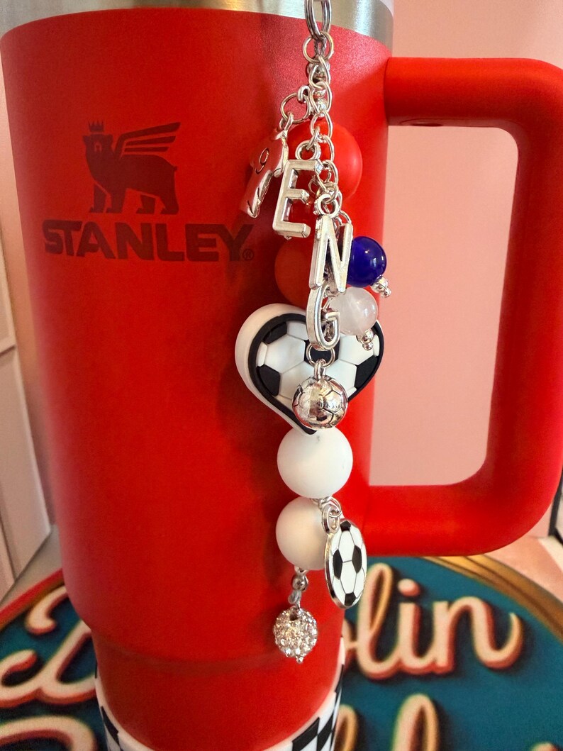 Puede incluir: Un vaso Stanley de color rojo brillante con asa tiene un llavero decorativo colgando de la parte superior. El llavero incluye dijes de letras plateadas que forman la palabra TENG, un dije de coraz&oacute;n con forma de bal&oacute;n de f&uacute;tbol, cuentas redondas blancas, naranjas y azules, y un peque&ntilde;o dije de bal&oacute;n de f&uacute;tbol.
