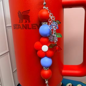 Op de afbeelding: Een rode Stanley-mok met een decoratieve bedel. De bedel heeft rode en blauwe kralen, een rode bloem en kleine bloemen- en steraccenten. De mok heeft een handvat en het Stanley-logo.