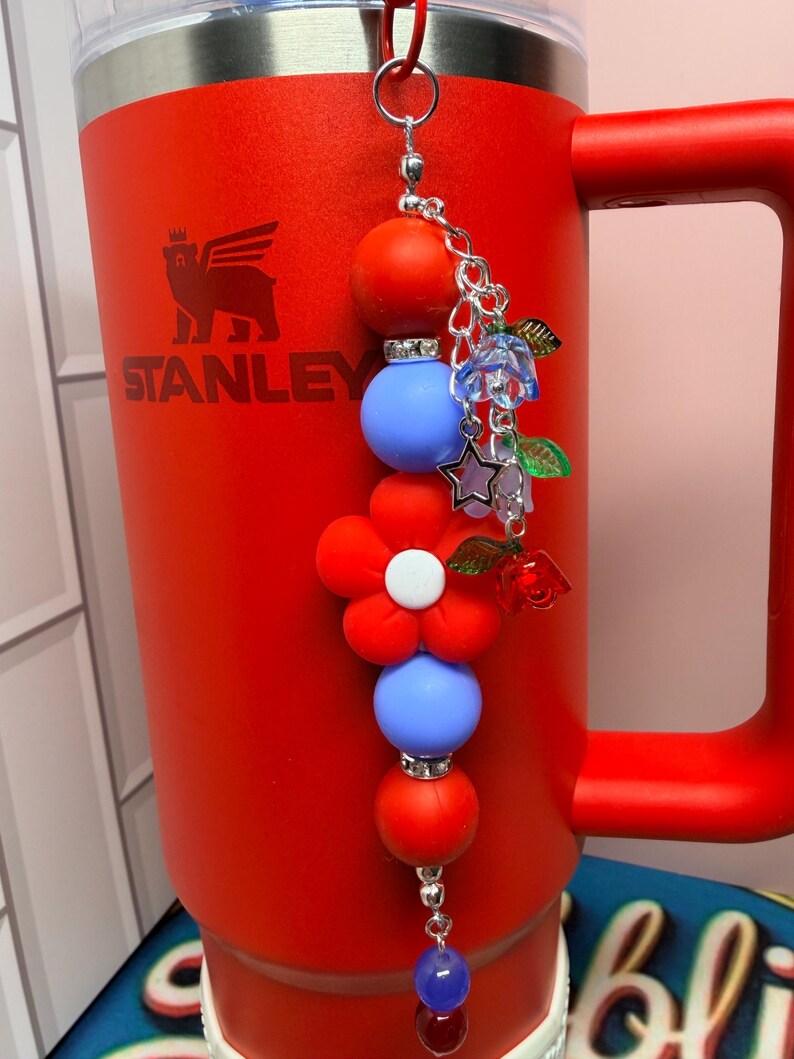 Op de afbeelding: Een rode Stanley-mok met een decoratieve bedel. De bedel heeft rode en blauwe kralen, een rode bloem en kleine bloemen- en steraccenten. De mok heeft een handvat en het Stanley-logo.