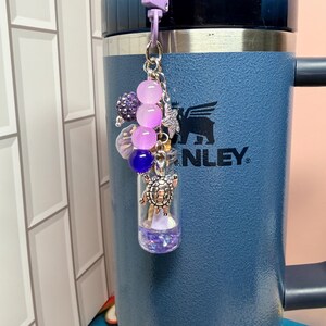 May include: A decorative keychain featuring a small clear glass vial filled with purple glitter, attached to a light purple lobster clasp. The charm includes a silver-toned turtle pendant, a small starfish, and a cluster of purple and blue beads.