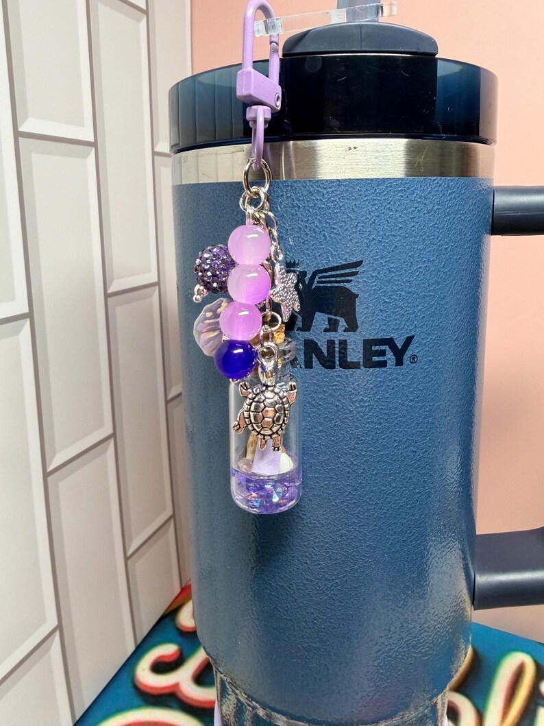 May include: A decorative keychain featuring a small clear glass vial filled with purple glitter, attached to a light purple lobster clasp. The charm includes a silver-toned turtle pendant, a small starfish, and a cluster of purple and blue beads.