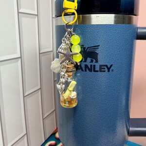 May include: A yellow lobster clasp keychain featuring a small glass bottle with a cork stopper, containing a rolled paper scroll and gold glitter. The charm includes a silver starfish, a mermaid figure, and three bright yellow translucent beads.