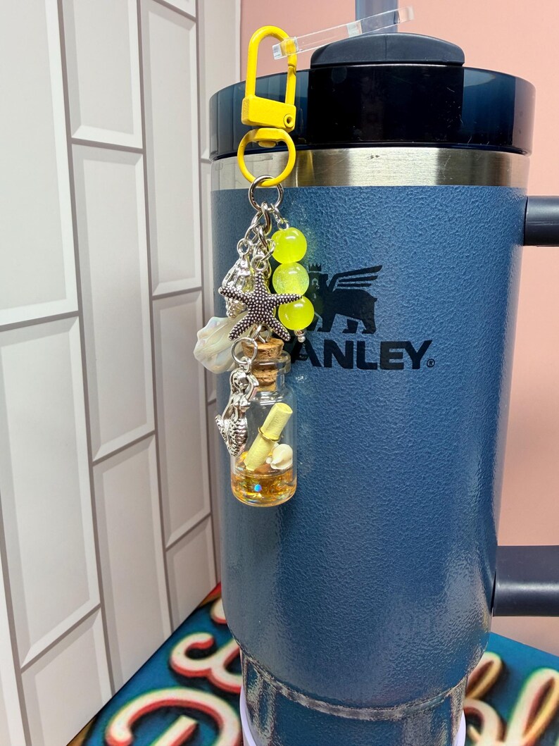 May include: A yellow lobster clasp keychain featuring a small glass bottle with a cork stopper, containing a rolled paper scroll and gold glitter. The charm includes a silver starfish, a mermaid figure, and three bright yellow translucent beads.
