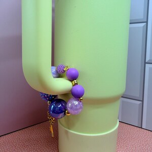 Op de afbeelding: Een kralenarmband met paarse, blauwe en transparante kralen hangt aan een lichtgroene beker. De armband heeft verschillende kralenmaten en texturen, waaronder een grote, iriserende blauwe kraal en kleinere, glinsterende paarse kralen.