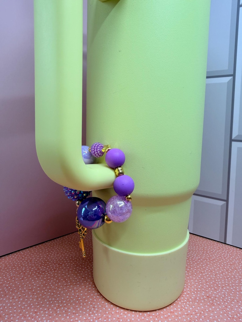Op de afbeelding: Een kralenarmband met paarse, blauwe en transparante kralen hangt aan een lichtgroene beker. De armband heeft verschillende kralenmaten en texturen, waaronder een grote, iriserende blauwe kraal en kleinere, glinsterende paarse kralen.