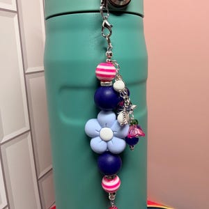 May include: A decorative beaded charm featuring a large light blue flower bead, dark blue round beads, and pink and white striped beads. The charm includes a silver-toned metal clasp, a small dragonfly charm, and a dangling pink textured bead.