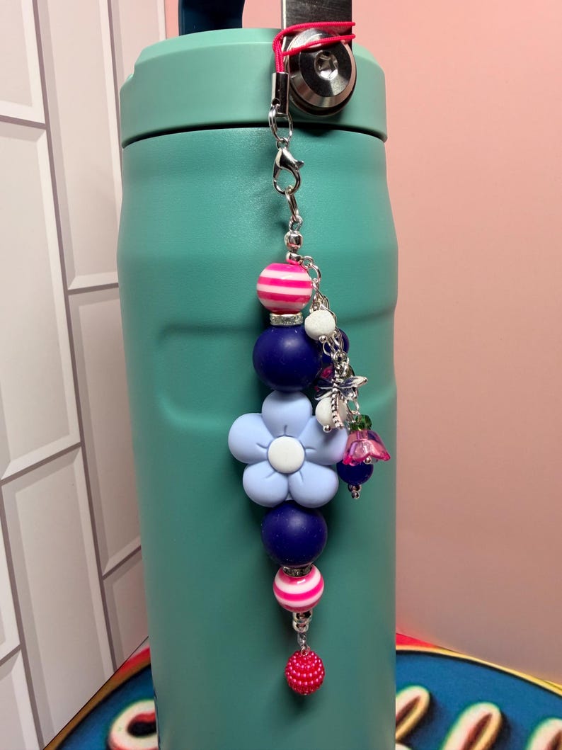 May include: A decorative beaded charm featuring a large light blue flower bead, dark blue round beads, and pink and white striped beads. The charm includes a silver-toned metal clasp, a small dragonfly charm, and a dangling pink textured bead.
