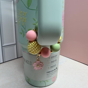 May include: A light green water bottle with floral designs and a beaded charm. The charm features pink, yellow, and green beads, and a pink flower pendant. The bottle has a handle and a white label with text.