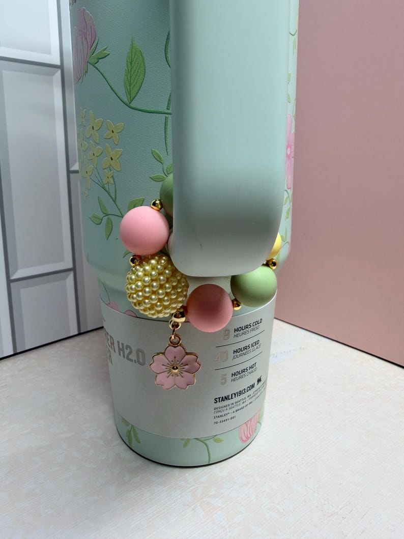 May include: A light green water bottle with floral designs and a beaded charm. The charm features pink, yellow, and green beads, and a pink flower pendant. The bottle has a handle and a white label with text.