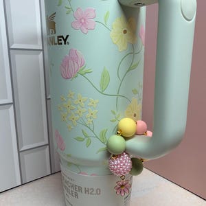 May include: A mint green Stanley Quencher H2.0 Flowstate tumbler with a floral design in pink and yellow. The tumbler has a handle and is accessorized with a beaded charm in pastel colors. The Stanley logo is visible.