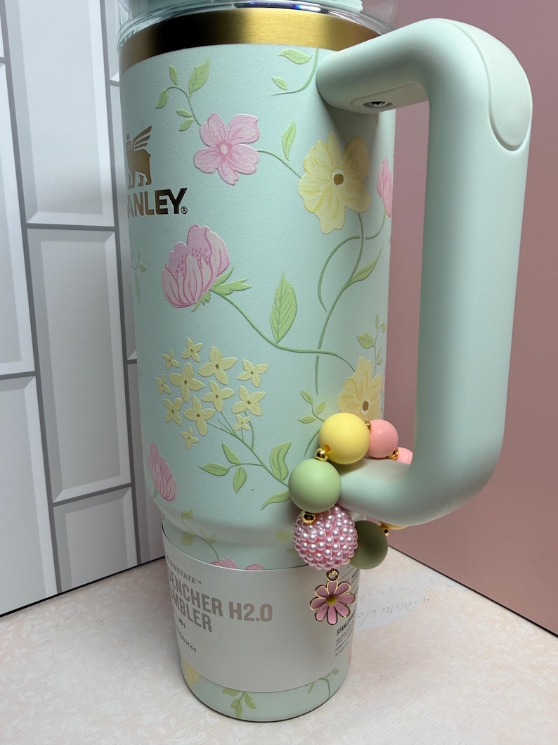 May include: A mint green Stanley Quencher H2.0 Flowstate tumbler with a floral design in pink and yellow. The tumbler has a handle and is accessorized with a beaded charm in pastel colors. The Stanley logo is visible.